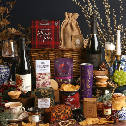 A rich Christmas hamper arrangement featuring items from the Winter Wonders Christmas Gift Basket, including a wicker basket, a box of mince pies, a bottle of Prosecco, a bottle of red wine, a tin of Yorkshire Crisps, a bar of Sticky Toffee Pudding chocolate, a purple bag of Cambrook nuts, a box of tea, and assorted treats, arranged on a dark table with candles, grapes, and decorative branches.