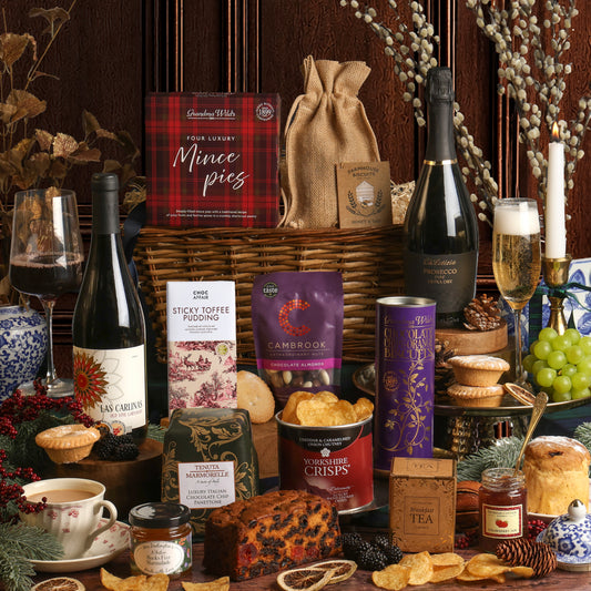 A rich Christmas hamper arrangement featuring items from the Winter Wonders Christmas Gift Basket, including a willow basket, a box of mince pies, a bottle of Prosecco, a bottle of red wine, a tin of Yorkshire Crisps, a bar of Sticky Toffee Pudding chocolate, a purple bag of Cambrook nuts, a box of tea, and assorted treats, arranged on a dark table with candles, grapes, and decorative branches.