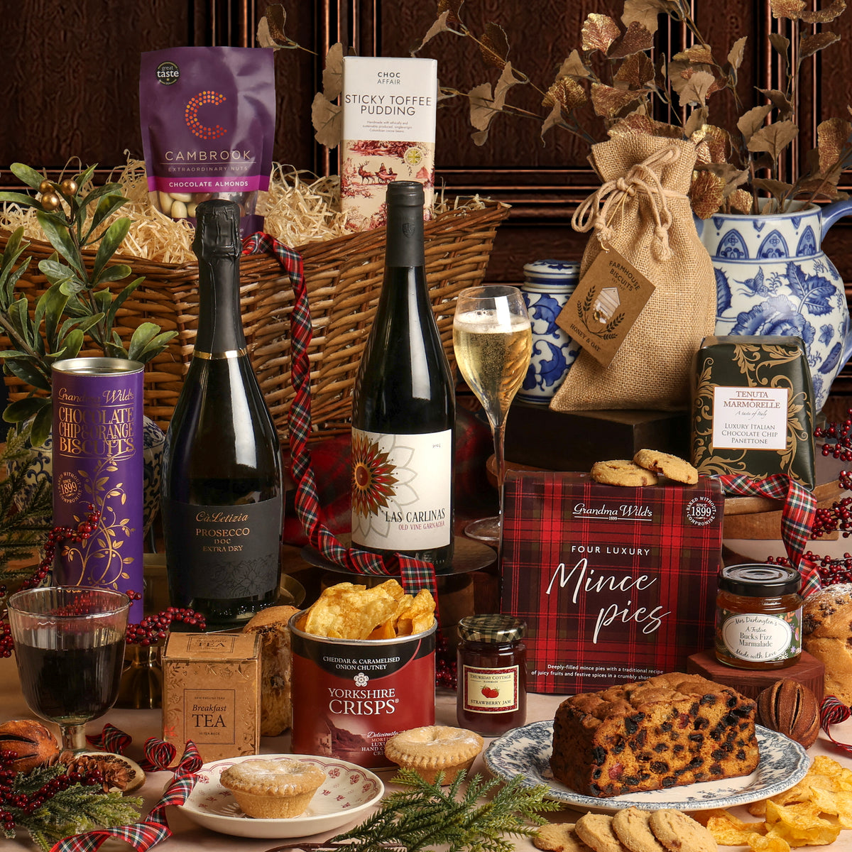 A festive Christmas hamper arrangement showing the Winter Wonders Christmas Basket, featuring a wine bottle, Prosecco, a glass of Prosecco, tea, mince pies, dried fruit, and crisps, styled with plaid ribbons and festive greenery in front of a willow basket.