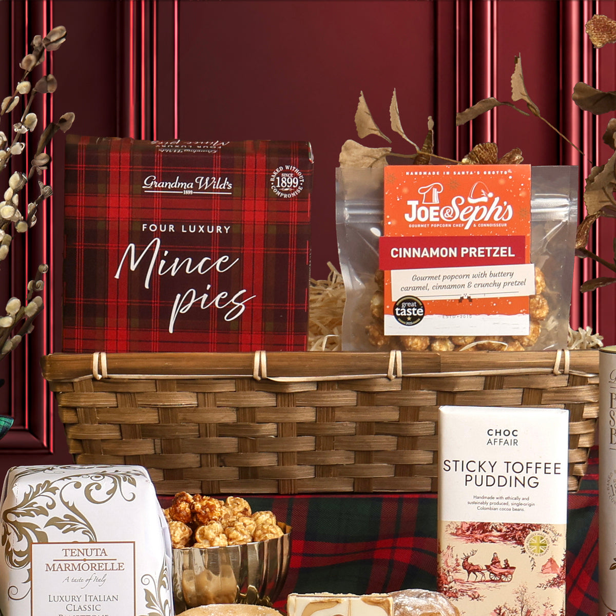 The White Christmas gift hamper arranged to show sweet food items alongside a bamboo basket, featuring a red plaid box of luxury mince pies, a bag of popcorn, a bar of chocolate, and other assorted treats. 