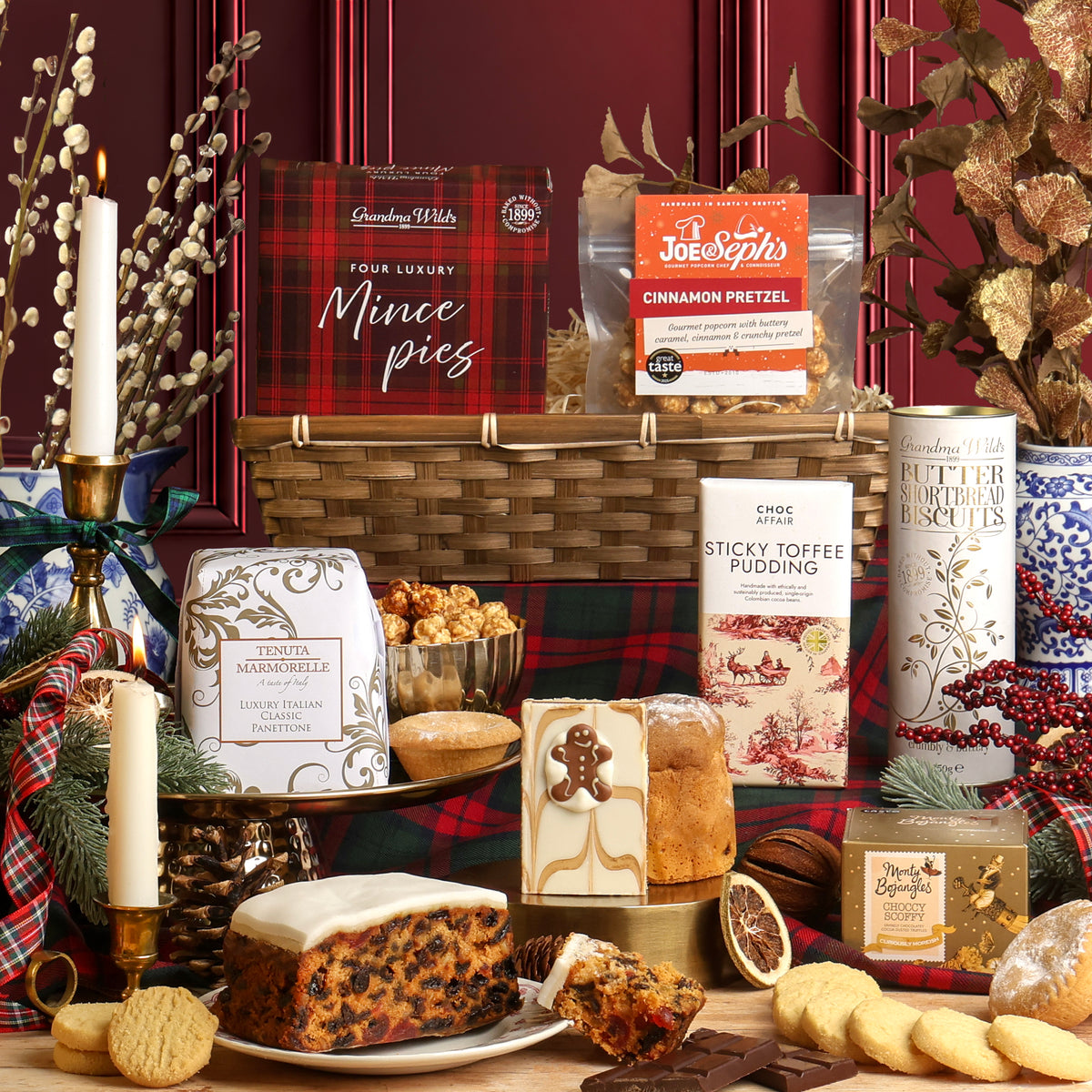 A White Christmas Gift Hamper with its contents displayed in front of the bamboo tray, arranged on a table with candles, plaid fabric, and festive decorations. Visible items include a bamboo tray, a box of mince pies, a bag of Joe & Seph's Cinnamon Pretzel popcorn, a tin of biscuits, a panettone, a Christmas fruit cake, and other treats.