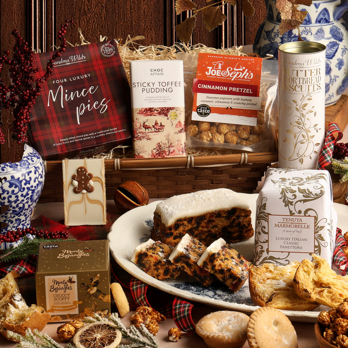The White Christmas hamper filled with assorted sweets and snacks, including a red plaid box of mince pies, a bar of sticky toffee pudding chocolate, a tin of butter shortbread biscuits, a box of Tenuta Marmorelle panettone, and a plate of sliced Christmas cake, arranged on a table with festive decorations.