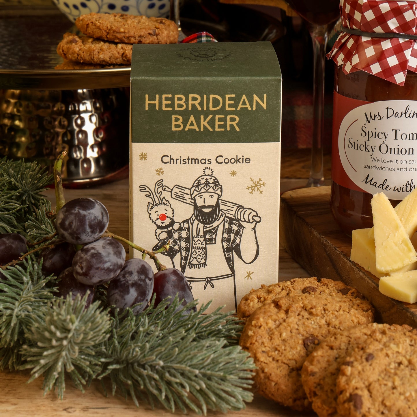 A Hebridean Baker Christmas Cookie box styled on a wooden table with cookies, grapes, greenery, and a jar of sticky onion chutney surrounding it, in a cosy Christmas kitchen setting.