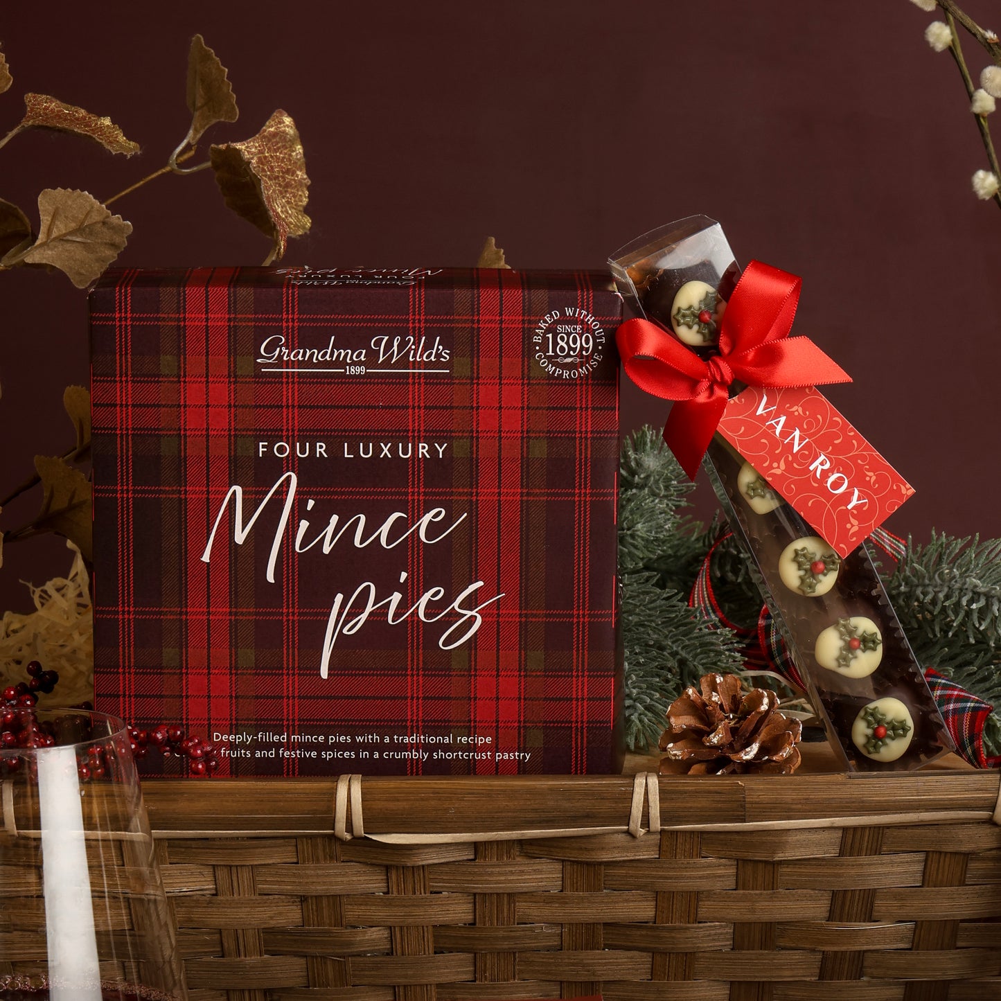 A scene showing contents from the Classic Christmas Food and Wine Hamper, suitable for couples to share, featuring a bamboo gift tray with a red and black plaid box of Grandma Wild's Four Luxury Mince pies inside, along with chocolate truffles decorated to look like Christmas puddings tied with a red ribbon and a Van Roy tag, set with festive decorations.