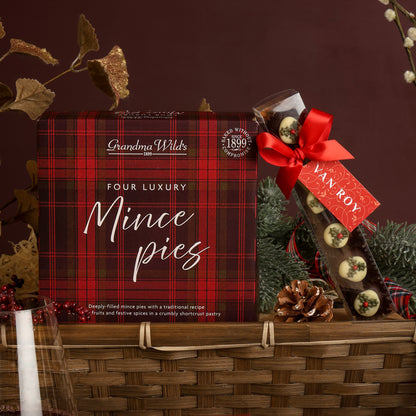 A scene showing contents from the Classic Christmas Food and Wine Hamper, suitable for couples to share, featuring a bamboo gift tray with a red and black plaid box of Grandma Wild's Four Luxury Mince pies inside, along with chocolate truffles decorated to look like Christmas puddings tied with a red ribbon and a Van Roy tag, set with festive decorations.