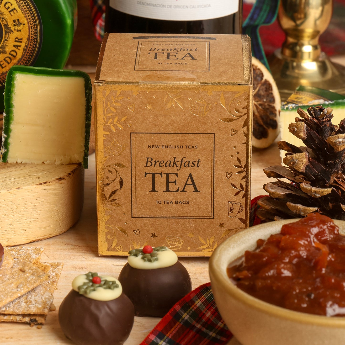 A small box of Breakfast Tea surrounded by Christmas props including a bowl of jam, chocolate treats, crackers, a wedge of cheese, and pinecones on a wooden surface.
