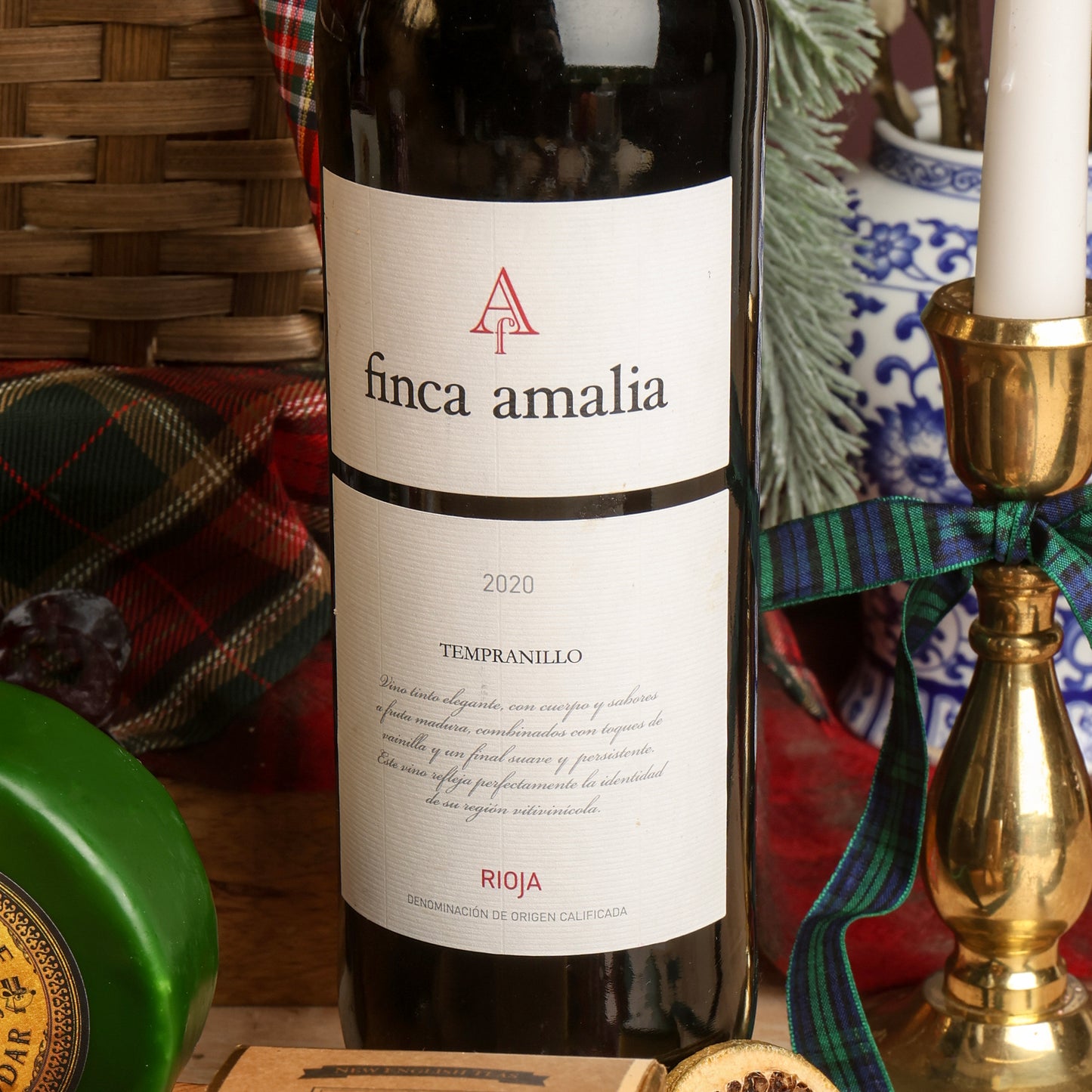 A bottle of Finca Amalia Rioja red wine, styled with a tartan plaid cloth, a green wheel of cheese, and a brass candlestick holder in a cosy setting.