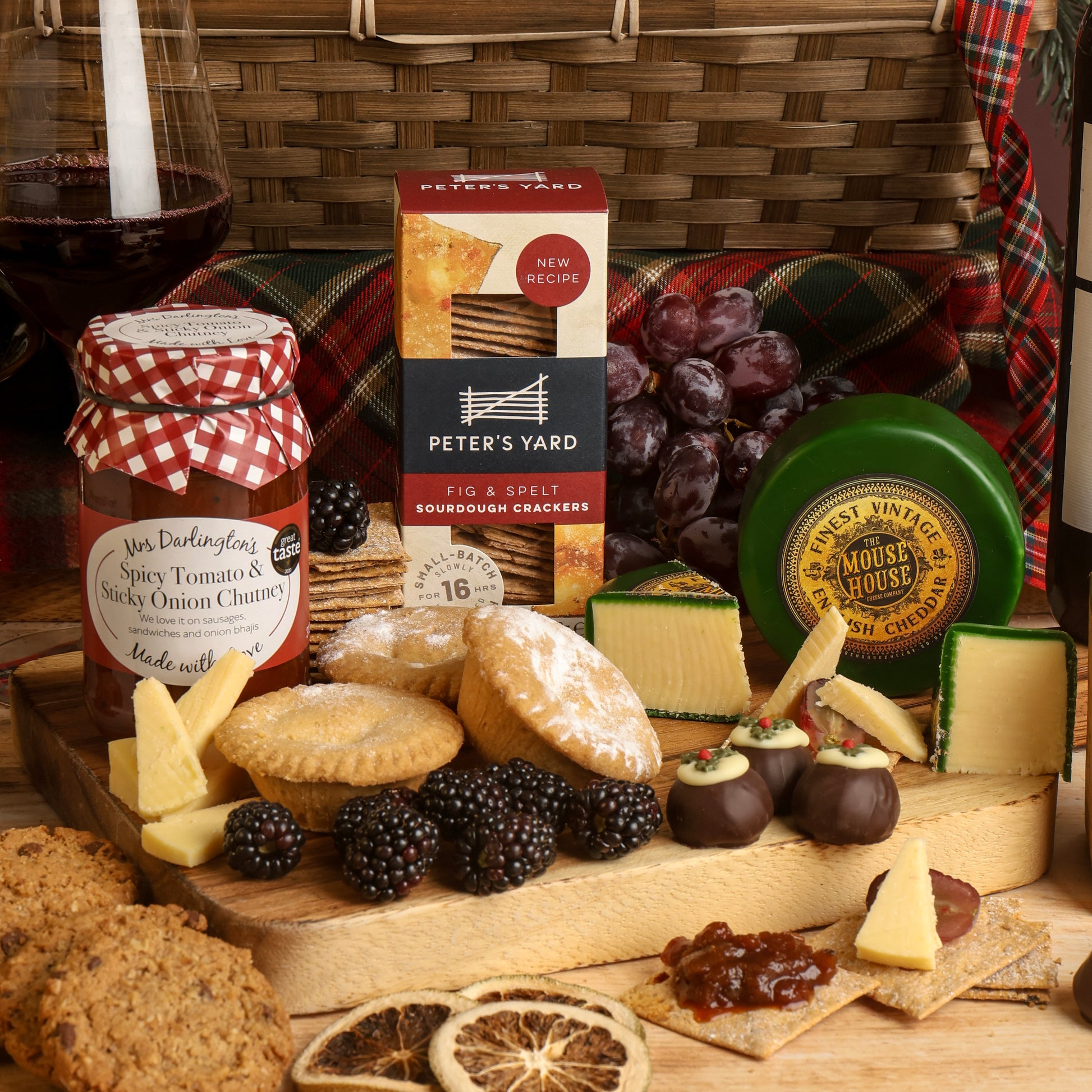 A collection of food and wine from the Classic Christmas Food and Wine Hamper, arranged on a wooden serving board, featuring a jar of Spicy Tomato & Sticky Onion Chutney, a box of Peter's Yard Sourdough Crackers, a green wheel of The Mouse House Cheddar cheese, cheese wedges, chocolates, a glass of red wine, with blackberries and dried orange slices to decorate the scene.