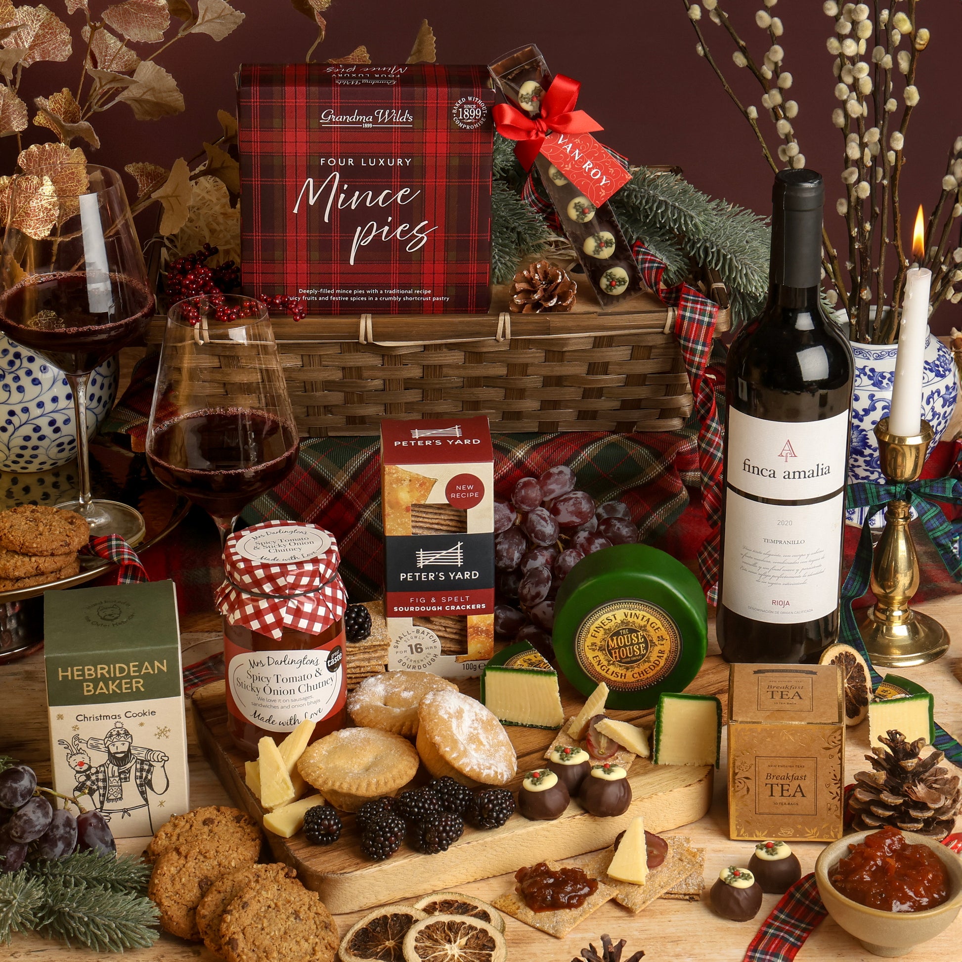 The Classic Christmas Food and Wine Hamper, with the contents displayed in front of the reusable bamboo tray, arranged on a table with a bottle of red wine and filled wine glasses, cheese and crackers, chocolates, tea, and packaged snacks. Visible products include Peter's Yard crackers, The Mouse House cheese, and a Hebridean Baker cookie.