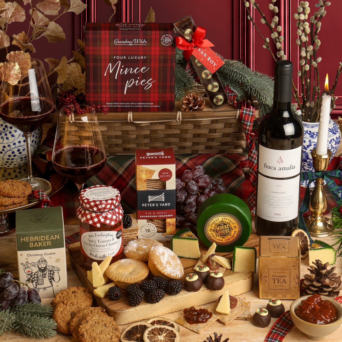 The Classic Christmas Food and Wine Hamper, with the contents displayed in front of the reusable bamboo tray, arranged on a table with a bottle of red wine and filled wine glasses, cheese and crackers, chocolates, tea, and packaged snacks. Visible products include Peter's Yard crackers, The Mouse House cheese, and a Hebridean Baker cookie.