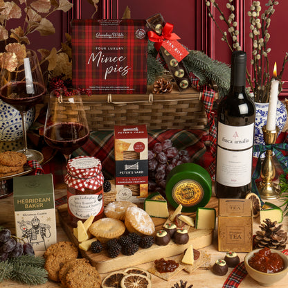 The Classic Christmas Food and Wine Hamper, with the contents displayed in front of the reusable bamboo tray, arranged on a table with a bottle of red wine and filled wine glasses, cheese and crackers, chocolates, tea, and packaged snacks. Visible products include Peter's Yard crackers, The Mouse House cheese, and a Hebridean Baker cookie.