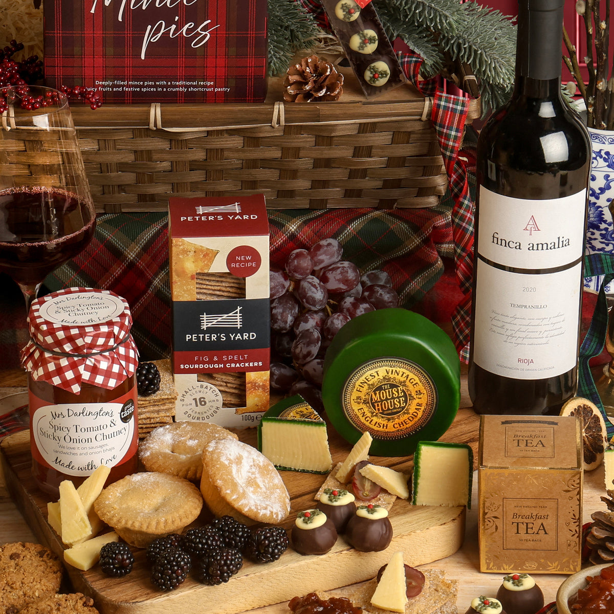 The Classic Christmas Food and Wine Hamper with charcuterie and cheese, a wine bottle, crackers and mince pies, styled with blackberries and grapes on a wooden board with red plaid fabric and festive decorations.