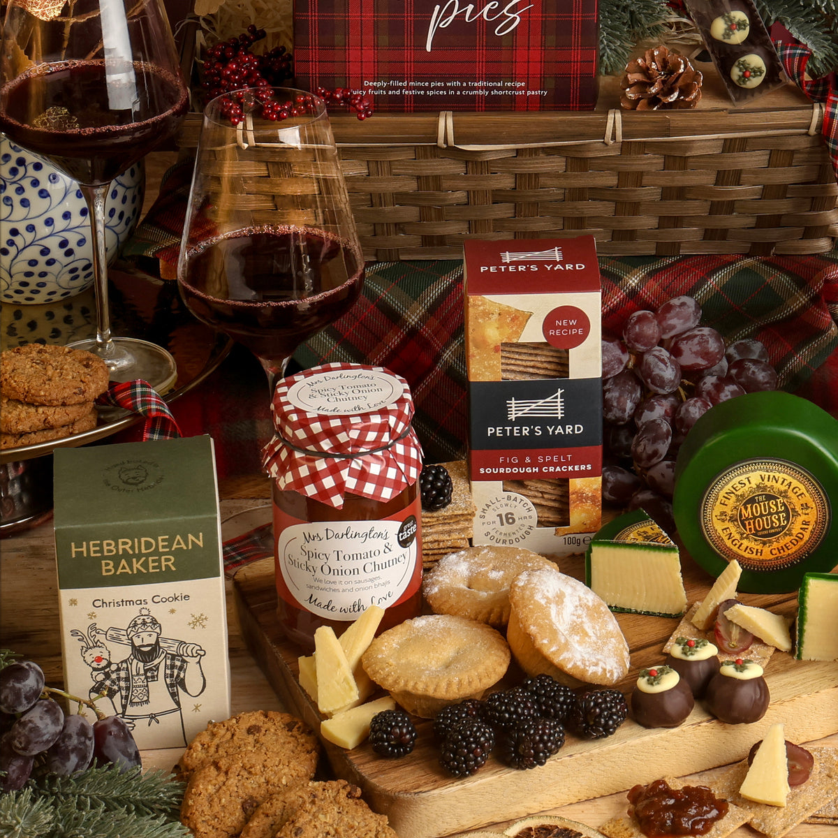 The Classic Christmas Food and Wine Hamper with festive food and drink, displayed around a bamboo basket, featuring sweet and savoury snacks, styled with cheese wedges, grapes, blackberries and wine glasses. 