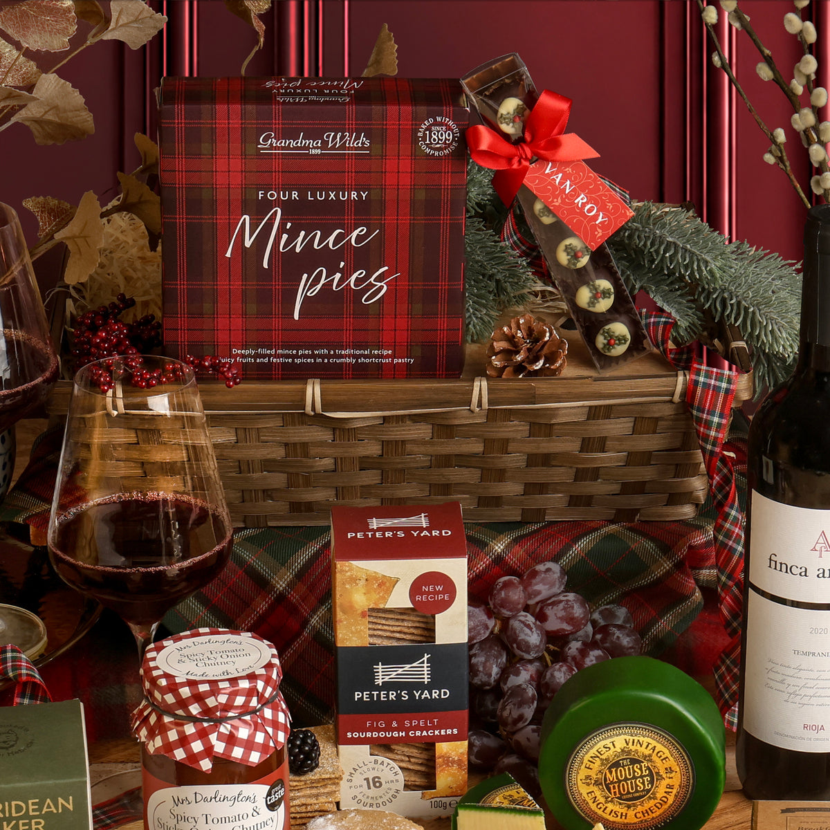 The Classic Christmas Food and Wine Hamper featuring a bamboo basket with a box of mince pies, Peter's Yard crackers, chocolate truffles, a jar of chutney, and cheese, set against a dark red background.