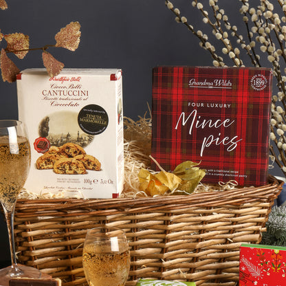 A willow gift basket with some of the contents from the Festive Prosecco Afternoon Tea Hamper stood upright inside, including a box of Cantuccini and a red plaid box of Grandma Wild's Four Luxury Mince Pies, styled with dried leaves and white branches with Prosecco glasses, against a dark grey background.