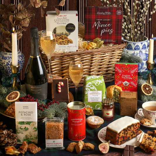 A Christmas assortment of food and drink from the Festive Prosecco Afternoon Tea Hamper, arranged on a table, including a bottle of Prosecco, a box of mince pies, a tin of biscuits, a bar of Pistachio and Rose chocolate, and other treats, with candles, pine branches, dried citrus, and a cup of tea.