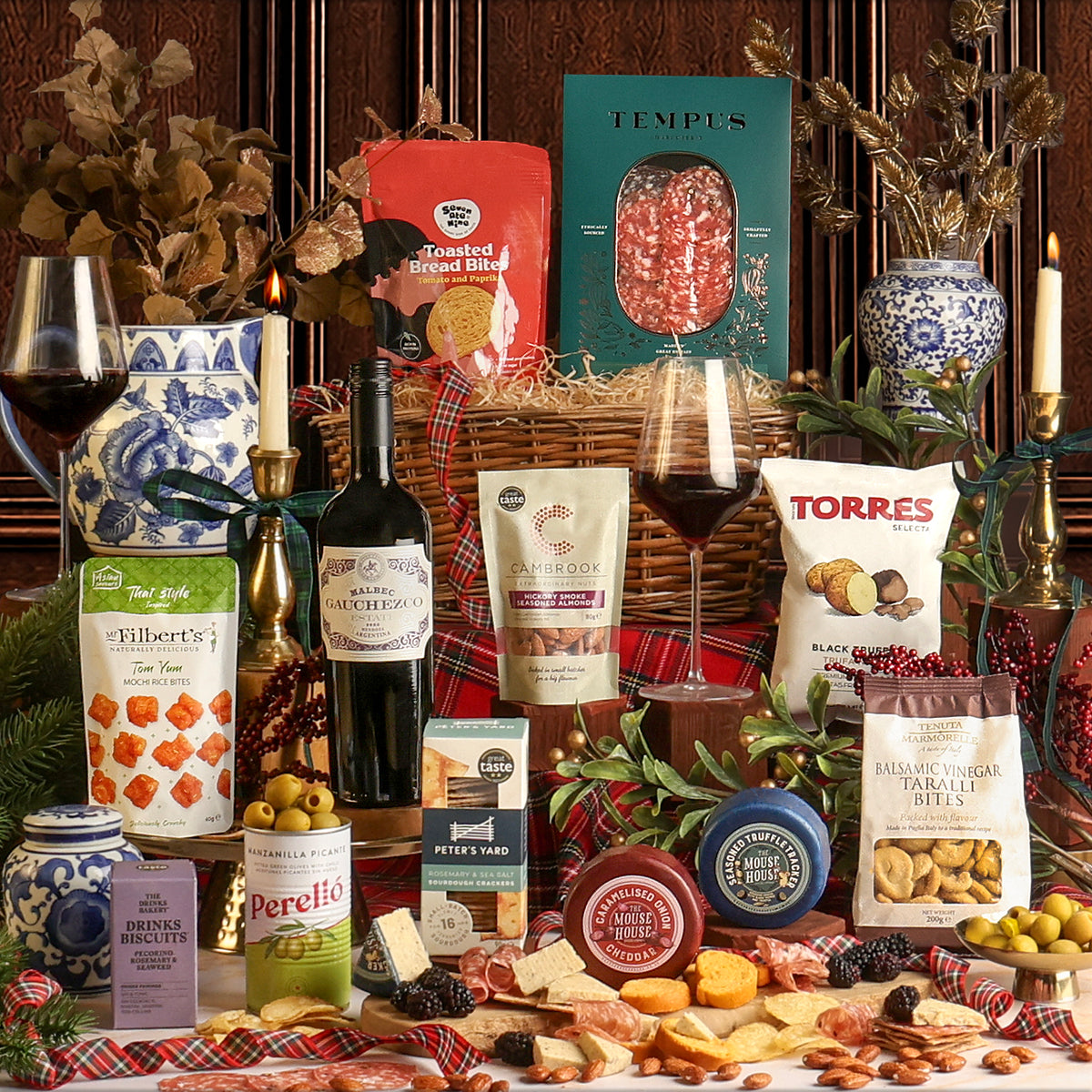 The Christmas Eve Wine and Nibbles Gift Basket, with assorted snacks, cheeses, crackers, wine, and packaged food items arranged around a wicker basket on a table with candles, glasses of red wine, and Christmas decor.