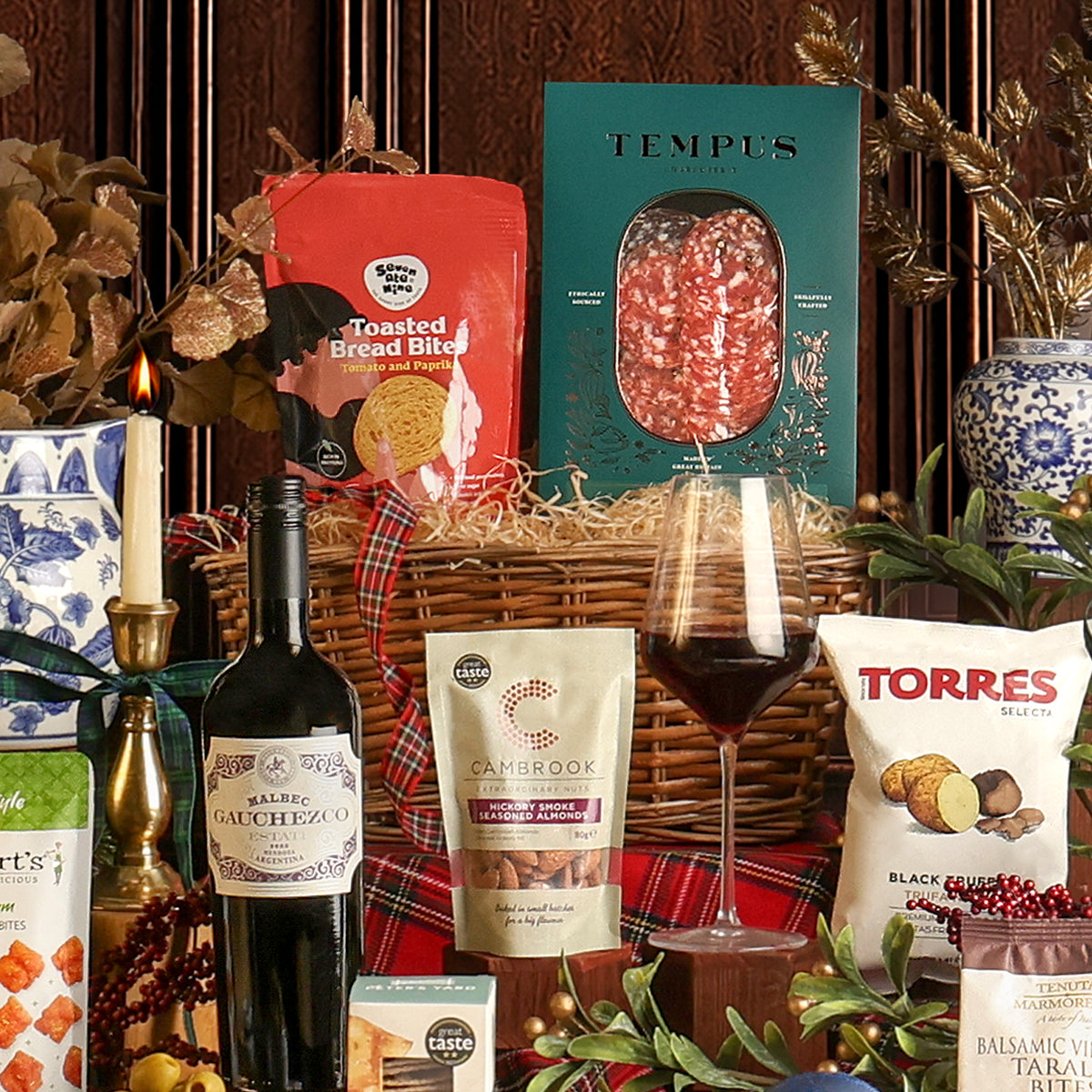 The Christmas Eve Wine and Nibbles gift basket contents arranged around a wicker basket with a red plaid ribbon, featuring a bottle of red wine, a pouch of Tempus charcuterie, Toasted Bread Bites, a bag of Cambrook nuts, a bag of Torres Black Truffle crisps, and a glass of red wine, surrounded by festive decorations.