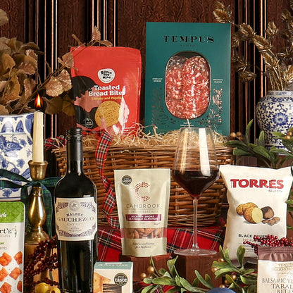 The Christmas Eve Wine and Nibbles gift basket contents arranged around a wicker basket with a red plaid ribbon, featuring a bottle of red wine, a pouch of Tempus charcuterie, Toasted Bread Bites, a bag of Cambrook nuts, a bag of Torres Black Truffle crisps, and a glass of red wine, surrounded by festive decorations.