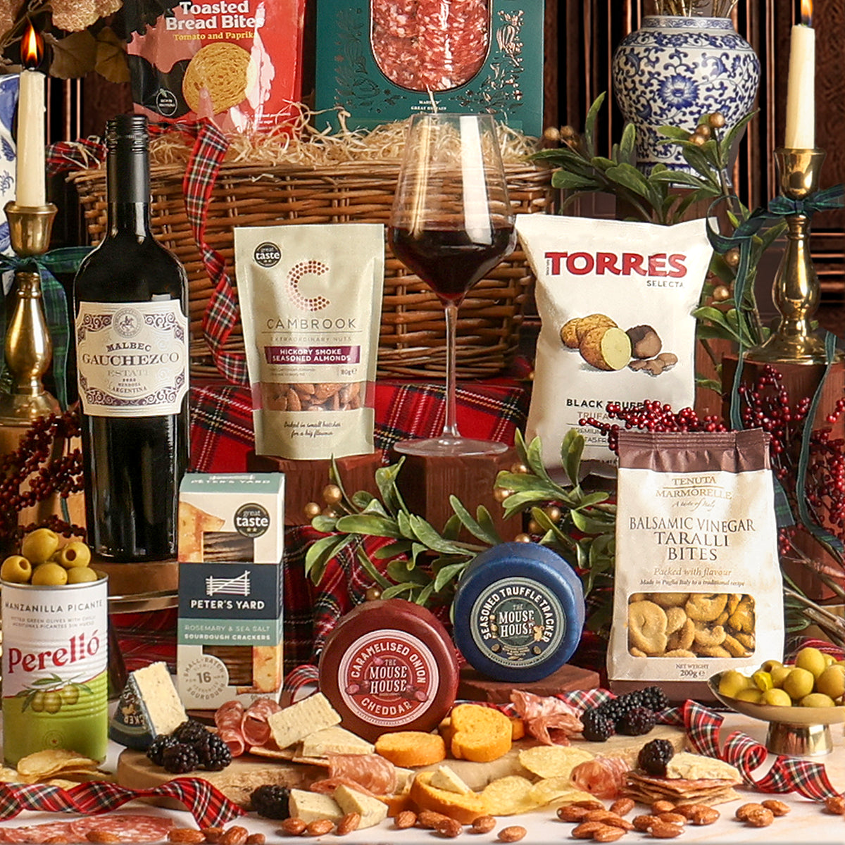A festive assortment of food and drink from the Christmas Eve Wine and Nibbles gift basket, with gourmet snacks, cheeses, crackers, and a bottle of red wine, styled with plaid fabric, candles, a glass of wine, and greenery.