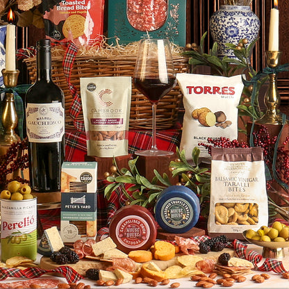 A festive assortment of food and drink from the Christmas Eve Wine and Nibbles gift basket, with gourmet snacks, cheeses, crackers, and a bottle of red wine, styled with plaid fabric, candles, a glass of wine, and greenery.