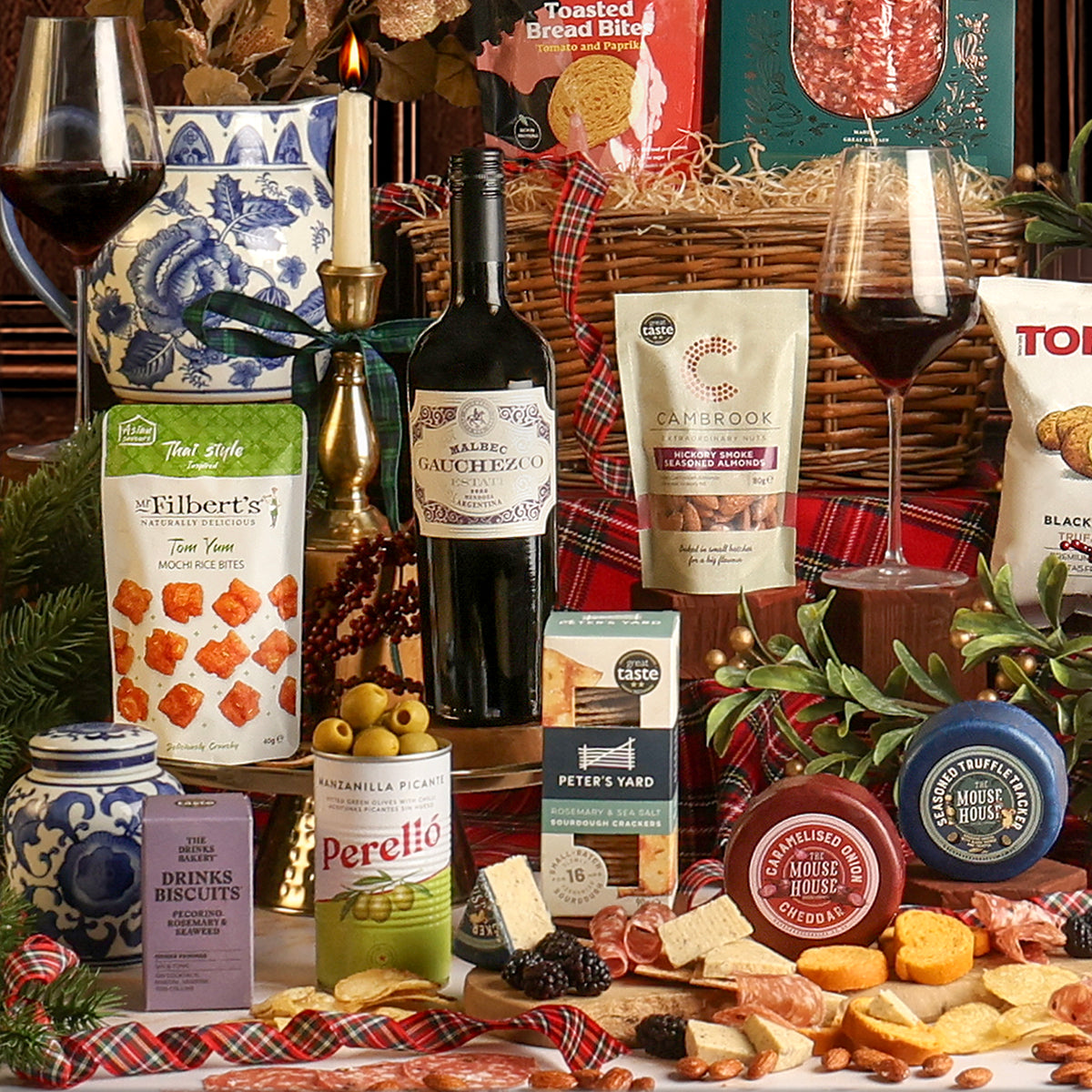 Food and drink from the Christmas Eve Wine and Nibbles gift basket, placed around the basket, including cheeses, crackers, wine, and snacks, styled with glasses of red wine, and festive decorations.
