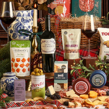 Food and drink from the Christmas Eve Wine and Nibbles gift basket, placed around the basket, including cheeses, crackers, wine, and snacks, styled with glasses of red wine, and festive decorations.