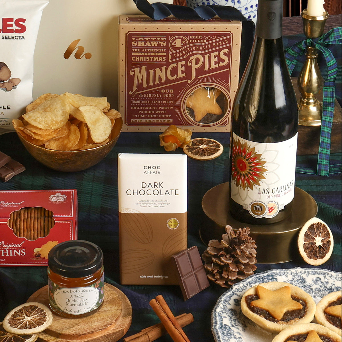 The Red Wine and Festive Treats Hamper, with its contents displayed on a plaid teal blanket, including a bottle of red wine, dark chocolate, crisps, marmalade and mince pies.