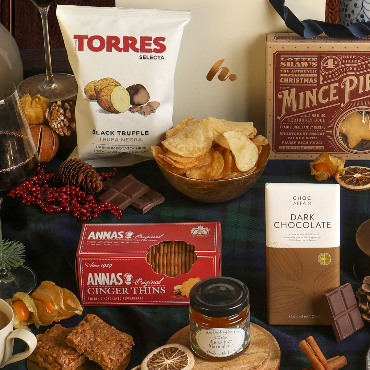 A styled festive gift hamper displaying some of the snacks included, featuring assorted chocolates, biscuits, and crisps on a dark green plaid fabric. 