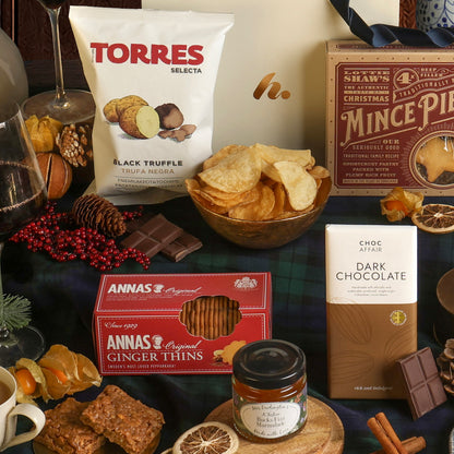 A styled festive gift hamper displaying some of the snacks included, featuring assorted chocolates, biscuits, and crisps on a dark green plaid fabric. 