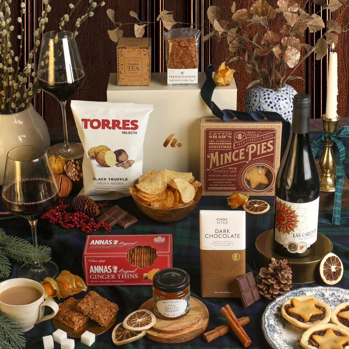 The vegan Red Wine and Festive Treats Hamper displayed to show the food and drink contents, arranged on a dark table with wine glasses, dried flowers, and candles for a Christmas theme. Visible products include Torres Black Truffle crisps, mince pies, ginger thins, a dark chocolate bar, and a bottle of red wine.