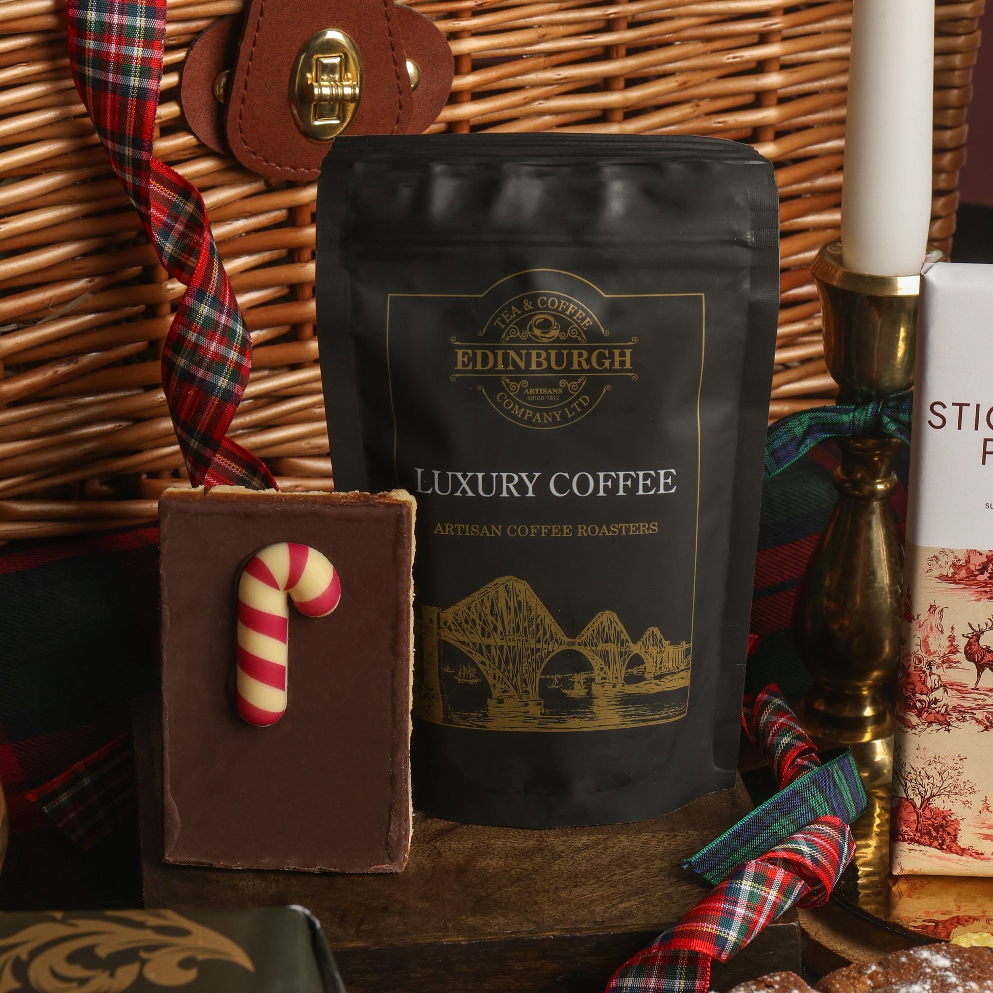 A black bag of Edinburgh Luxury Coffee stood upright on a wooden surface beside a chocolate flapjack with a candy cane decoration, with a wicker basket in the background, and a candlestick tied with plaid ribbon.