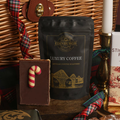 A black bag of Edinburgh Luxury Coffee stood upright on a wooden surface beside a chocolate flapjack with a candy cane decoration, with a wicker basket in the background, and a candlestick tied with plaid ribbon.