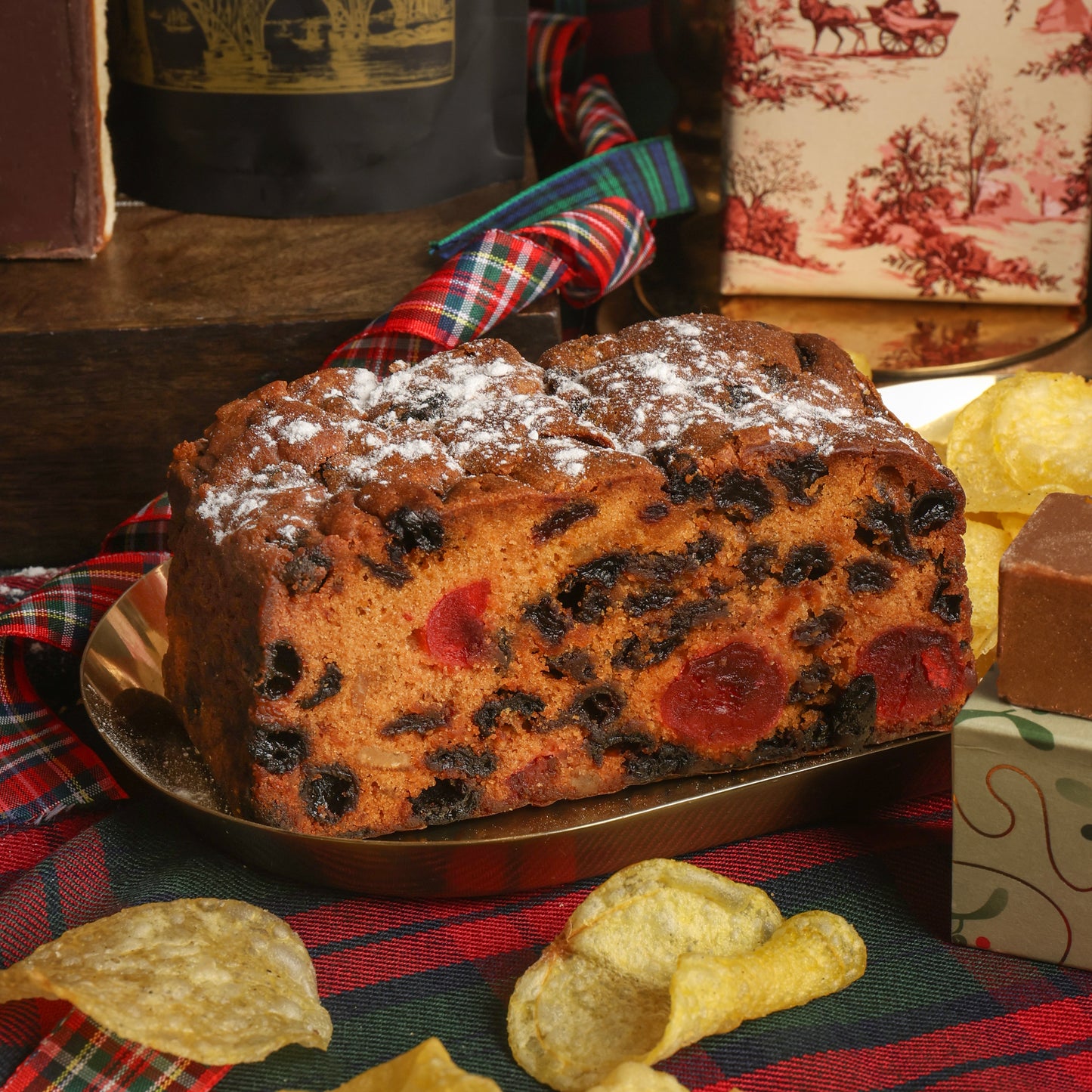 A Christmas fruit cake with fruit pieces, dusted with powdered sugar and served on a plate with crisps scattered in front, on a plaid tablecloth.
