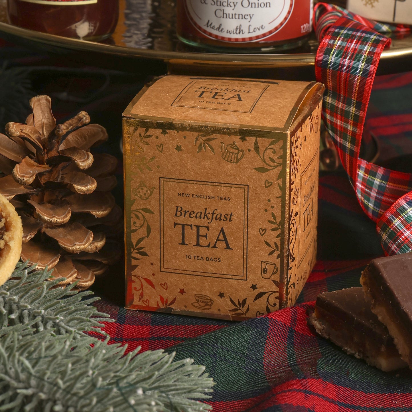 A paper box filled with breakfast tea on a red and green plaid fabric cloth with Christmas props including a pinecone, pine branches, a ribbon, and snacks in the background.