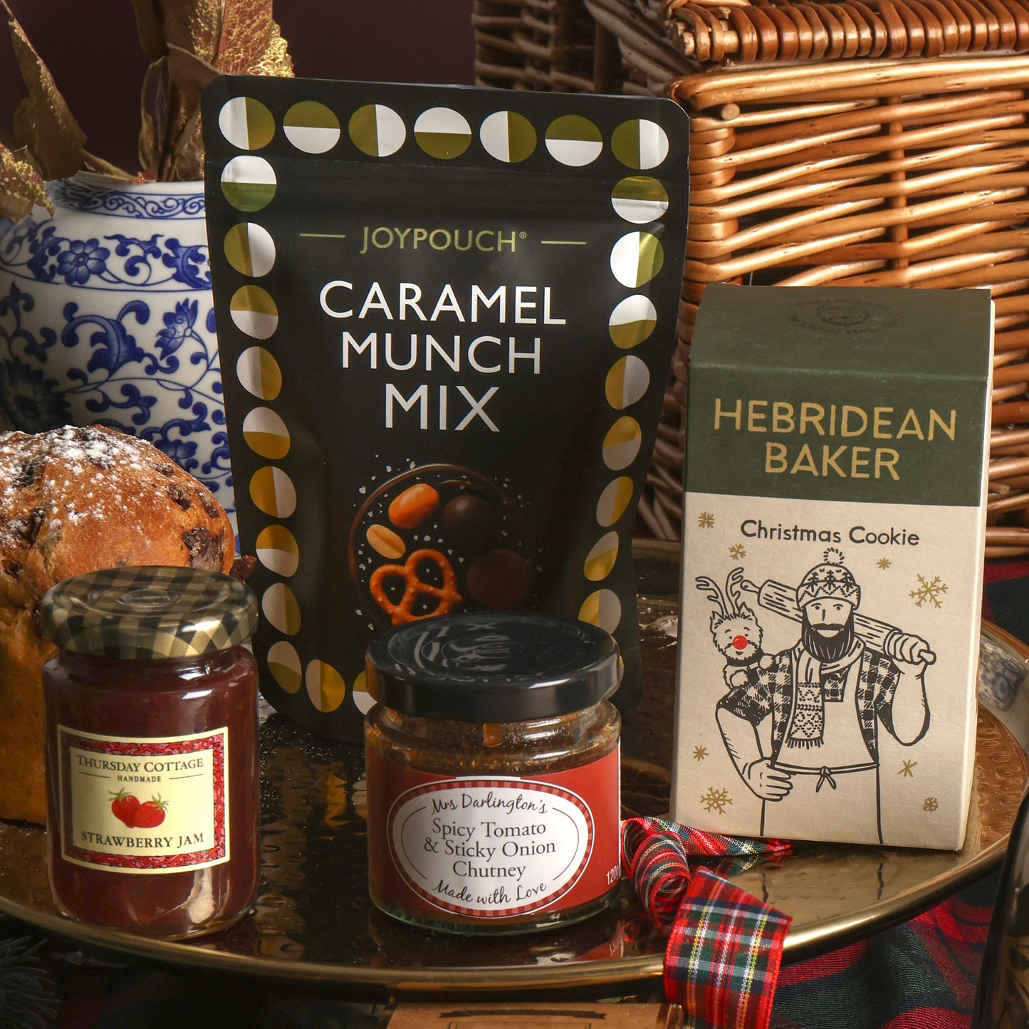 A Christmas wicker hamper with some of its food contents assorted on a gold tray in front, featuring a bag of nuts and pretzels, a boxed Hebridean Baker Christmas Cookie, a jar of Strawberry Jam, and a jar of Spicy Tomato & Sticky Onion Chutney, with a plaid ribbon.
