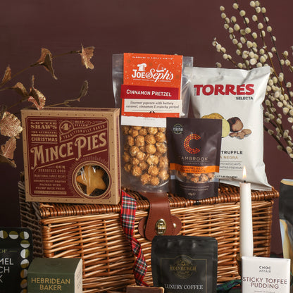 The wicker hamper and contents from The Magic of Christmas Hamper, displaying food around the hamper, including mince pies, Joe & Seph's Cinnamon Pretzel popcorn, Torres Truffle Crisps, and a bag of Cambrook nuts, set against a dark brown background with decorative dried branches.