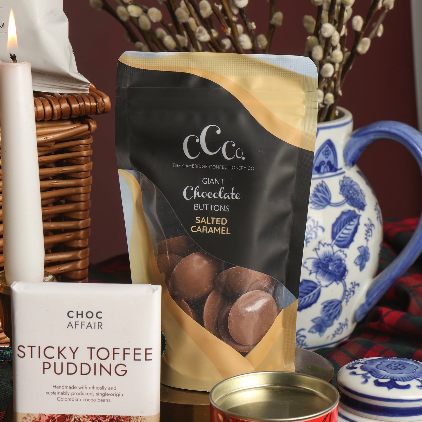 A styled product display featuring a bag of The Cambridge Confectionary Company Giant Chocolate Buttons, next to a bar of Choc Affair Sticky Toffee Pudding chocolate, with a lit candle, wicker basket, and blue-and-white ceramic jug in the background to decorate the scene.