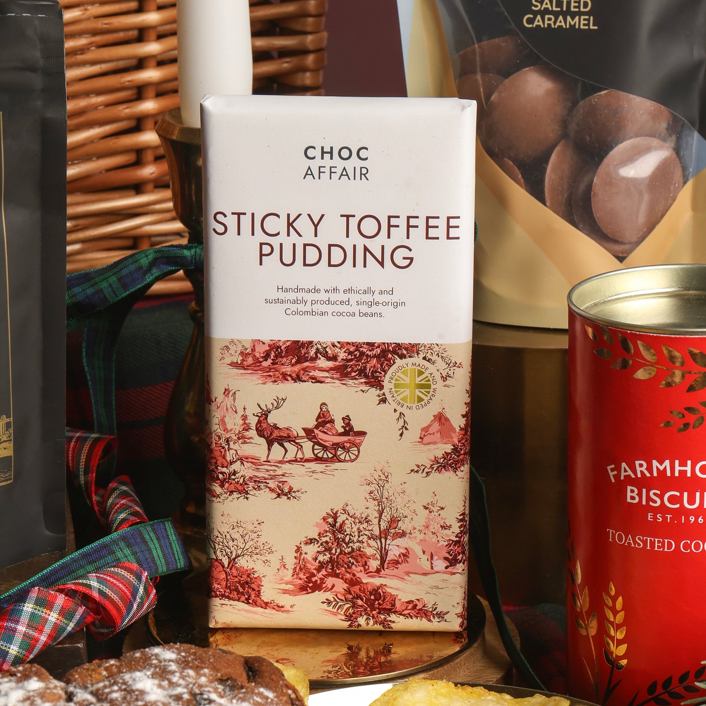 A bar of Choc Affair Sticky Toffee Pudding chocolate stands in a styled gift setting with other food items around it, including a red tin of biscuits and a bag of salted caramel chocolate buttons.