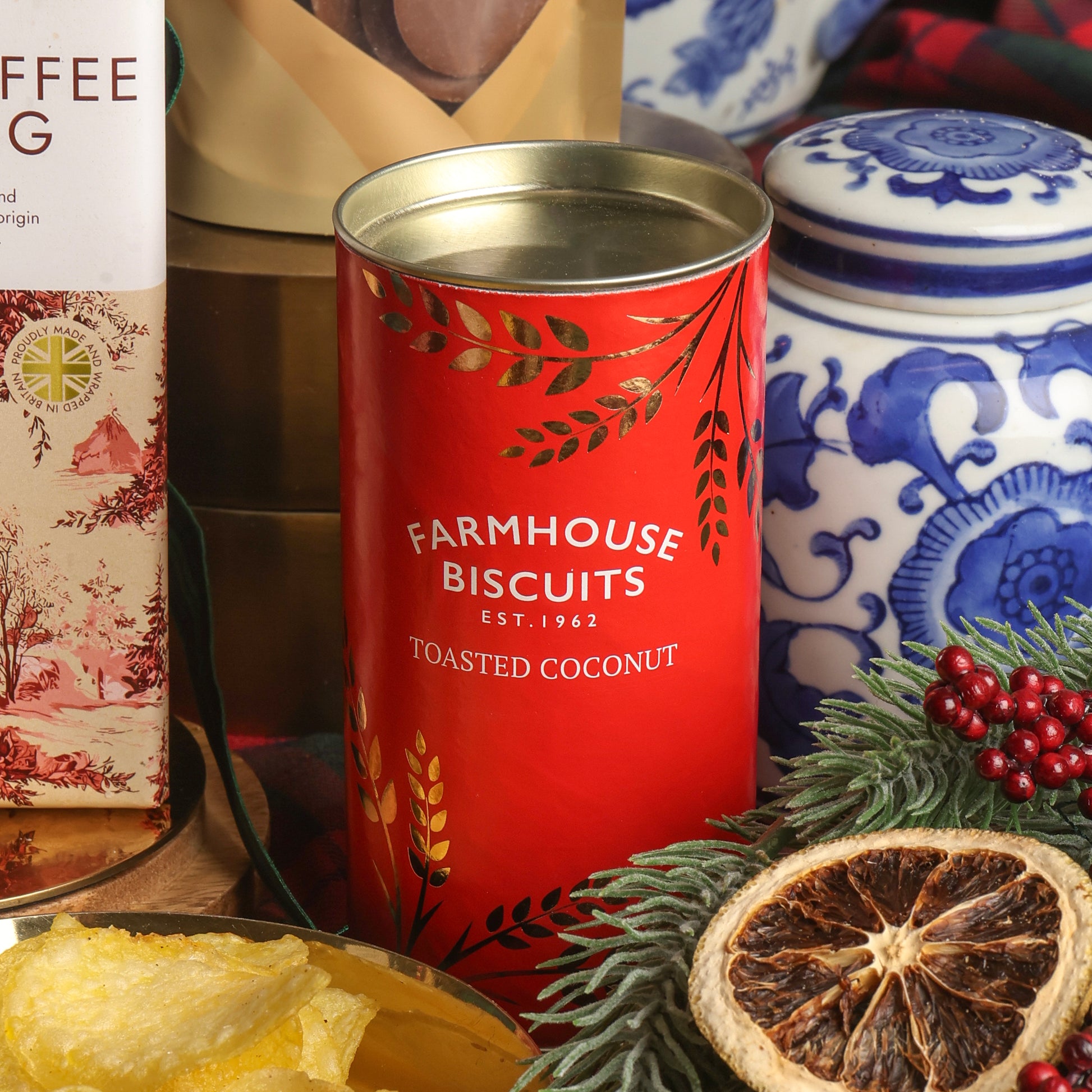 A red cylindrical tin of Toasted Coconut Farmhouse Biscuits in the centre, surrounded by Christmas props including a sliced dried orange, greenery, red berries, and a blue-and-white ceramic jar in the background.