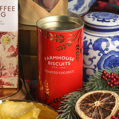 A red cylindrical tin of Toasted Coconut Farmhouse Biscuits in the centre, surrounded by Christmas props including a sliced dried orange, greenery, red berries, and a blue-and-white ceramic jar in the background.
