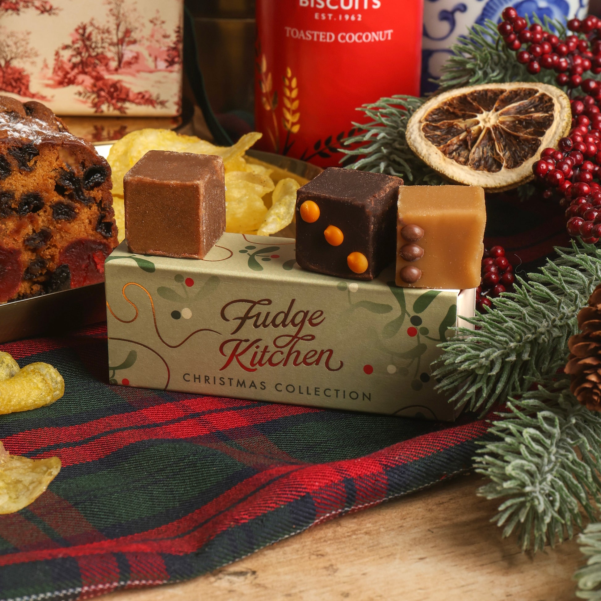 Three decorated fudge pieces displayed on top of a Fudge Kitchen Christmas Collection box, surrounded by festive decorations including pine branches, a dried orange slice, red berries, and a plaid cloth.
