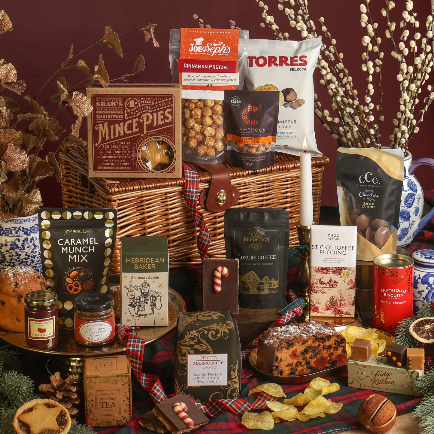 The Magic of Christmas Hamper displayed to show the contents in front of the wicker hamper, including mince pies, caramel munch mix, Hebridean Baker Christmas cookie, tea, coffee, and a fruit cake, with more snacks, arranged on a plaid blanket with seasonal decorations.