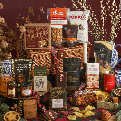 The Magic of Christmas Hamper displayed to show the contents in front of the wicker hamper, including mince pies, caramel munch mix, Hebridean Baker Christmas cookie, tea, coffee, and a fruit cake, with more snacks, arranged on a plaid blanket with seasonal decorations.