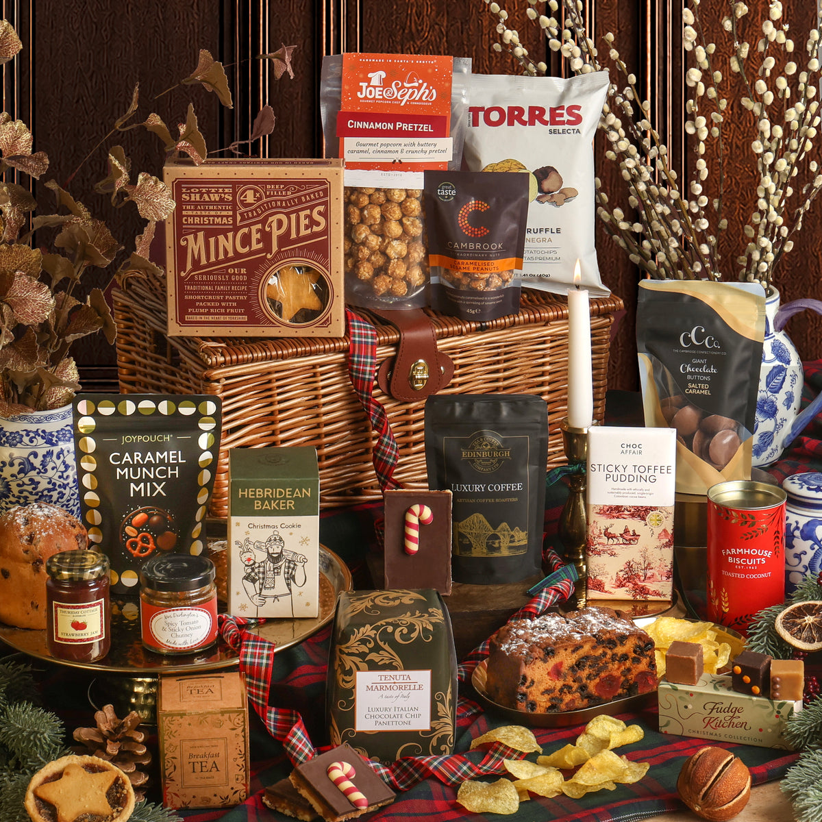 The Magic of Christmas Hamper displayed to show the contents in front of the wicker hamper, including mince pies, caramel munch mix, Hebridean Baker Christmas cookie, tea, coffee, and a fruit cake, with more snacks, arranged on a plaid blanket with seasonal decorations.