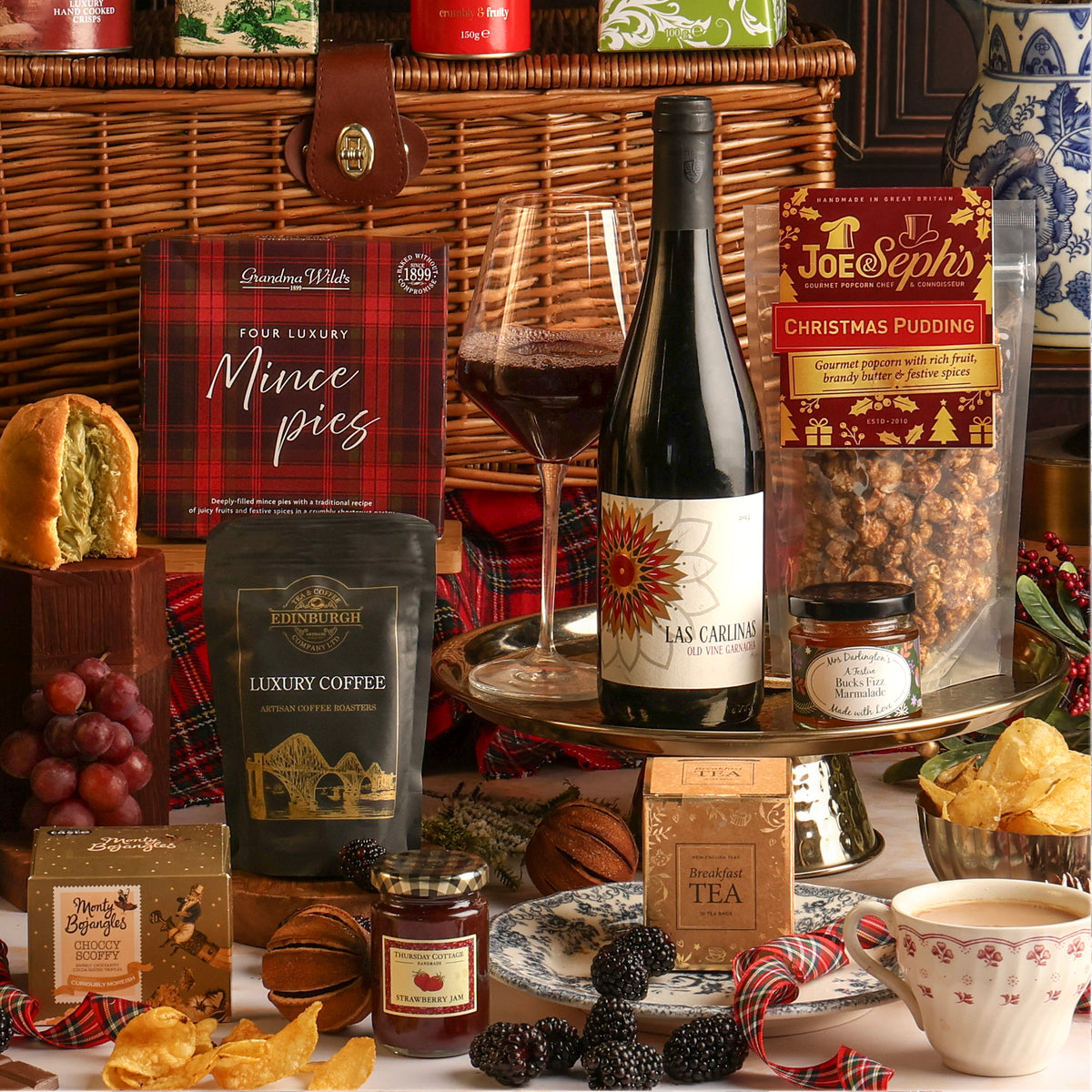 A luxury Christmas hamper gift set arranged on a table in front of a basket. The set includes a bottle of Las Carlinas red wine, a bag of coffee, a box of mince pies, a bag of Joe & Seph's Christmas Pudding popcorn, a jar of jam, a box of tea, styled with a cup of tea, grapes, blackberries, crisps, and other festive treats.