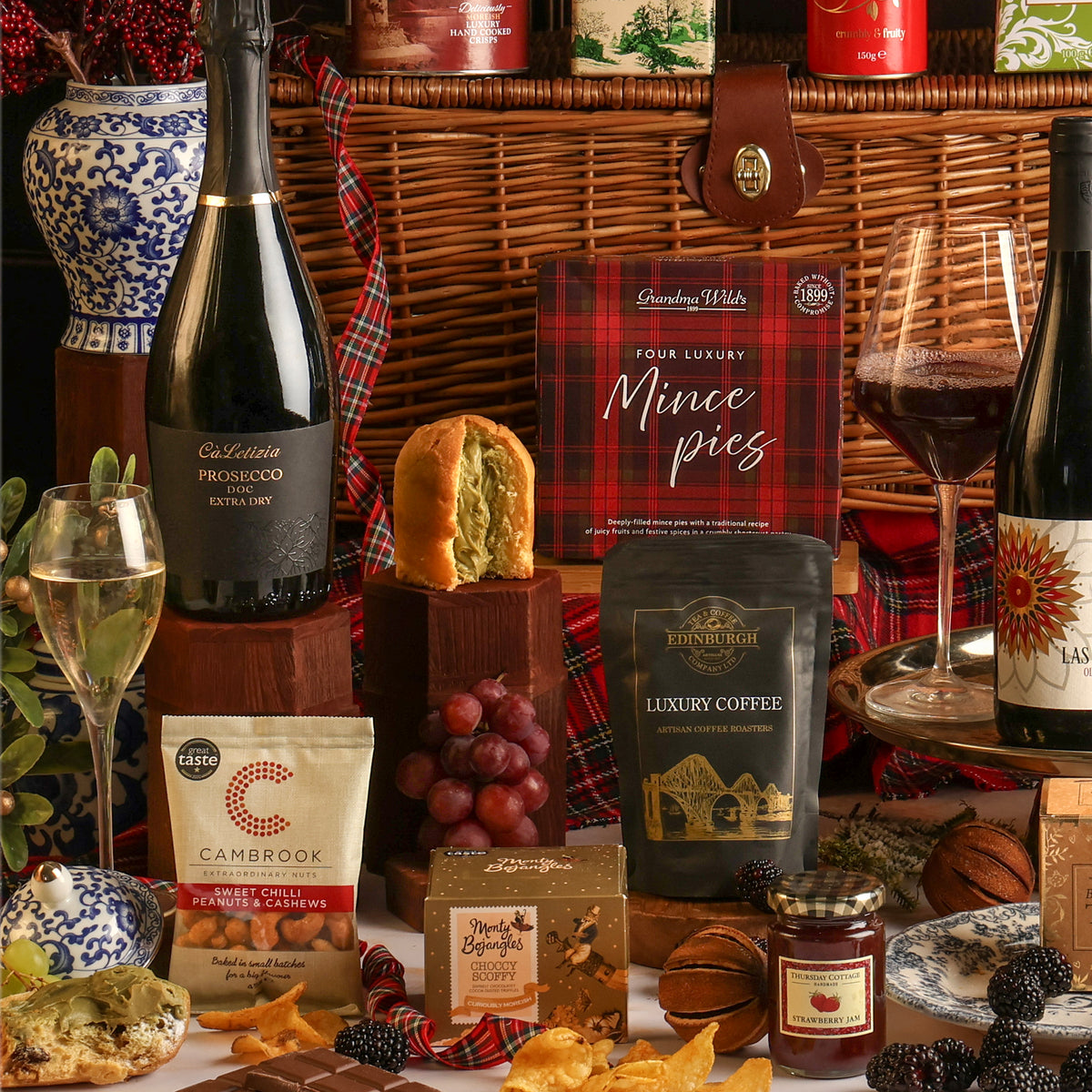 A luxury gift hamper filled with assorted treats, including a bottle of Prosecco, a bottle of red wine, a panettone, mince pies, a bag of Cambrook nuts, a jar of jam, tea and coffee, arranged in front of a basket.