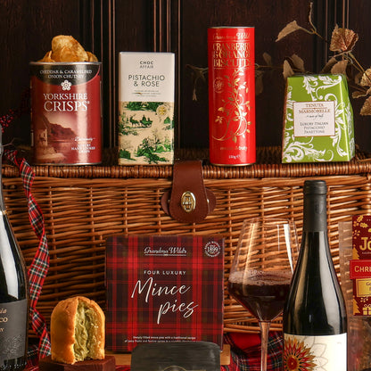A luxury hamper filled with gourmet treats, paired with a bottle of red wine, styled with a glass of red wine, a panettone, and ribbon, arranged against a dark wooden background. Visible items include Yorkshire Crisps, Pistachio & Rose chocolate, biscuits, and mince pies.