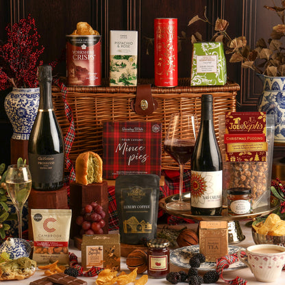 A Luxury Bearing Gifts Christmas hamper gift set arranged with a wicker basket, featuring wine and Prosecco bottles, boxed treats, tea, coffee, crisps, and seasonal decorations on a dark wood background.