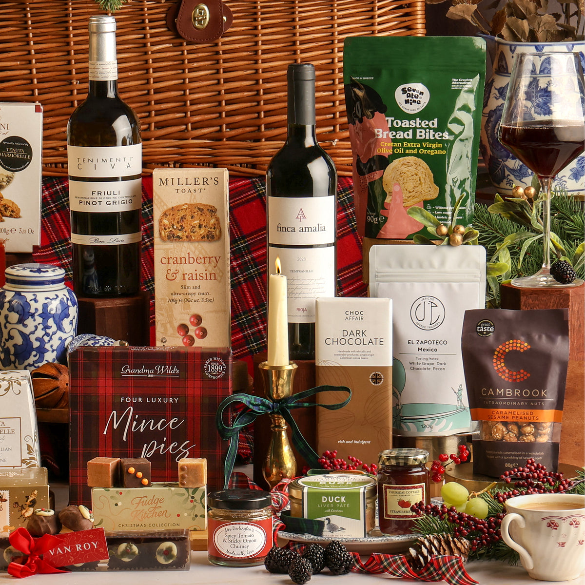 A festive gift hamper arrangement featuring bottles of wine, a bag of toasted bread bites, mince pies, chocolates, and tea, styled with a candle, plaid fabric, pine branches, and Christmas decorations.